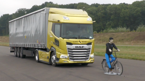 A lorry with a golden cab and grey trailer drives towards a life-sized model of a cyclist, it looks like both are on a flat concrete surface resembling a runway.