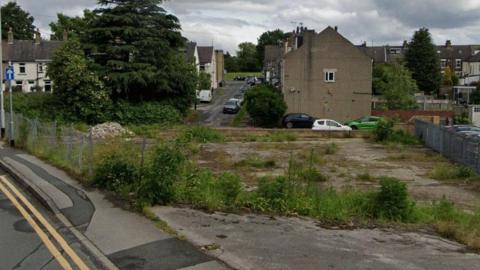 A derelict plot of land with weeds growing out of the concrete. On one side is a pavement and road. Behind the plot are rows of terraced houses with cars parked along the roads.