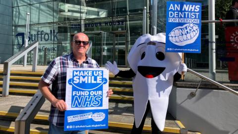 A bald man with sunglasses and a chequered shirt standing outside a glass building next to someone dressed as a tooth. Both are holding up placards calling for more NHS dentists