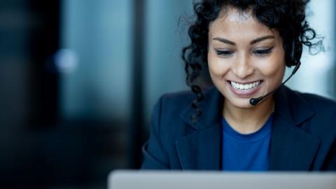 A woman wearing a blue t-shirt and a navy blazer looks down towards a laptop while smiling. She is wearing headphones and a microphone that wraps around the side of her face. 