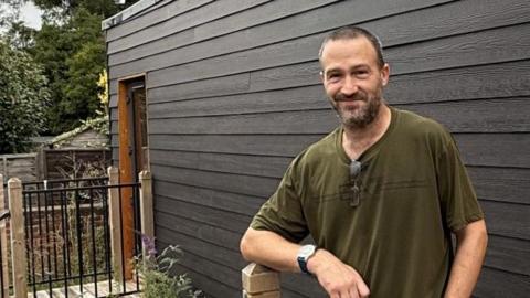 A smiling man with a green T-shirt and black trousers standing in front of a black and wooden pod for the homeless where he now lives