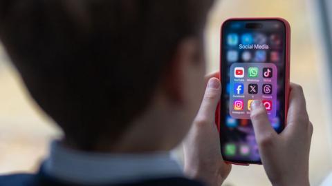 A 12-year-old boy looks at an iPhone screen showing various social media apps including TikTok, Facebook and X 