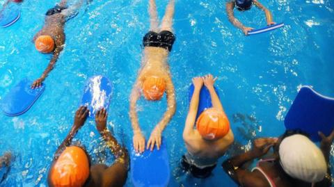 Children in a pool taking part in a swimming lesson. They are swimming on their fronts holding out blue floats, while adults stand in the water watching