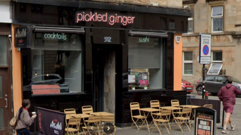 A Glasgow restaurant, sitting on the corner of a road. Several wooden tabls and chairs are set up outside, while the name - Pickled Ginger - is in orange or red lighting above the entrance. 