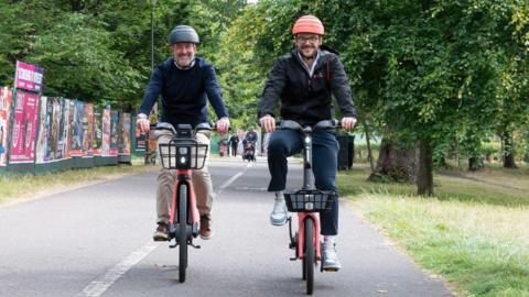 Transport and Environment Convener,Stephen Jenkinson (left) wearing a dark jumper and light trousers and Voi UK General Manager, James Bolton (right) wearing dark clothing test out the new e-bikes at the launch of Edinburgh's trial cycle hire scheme.