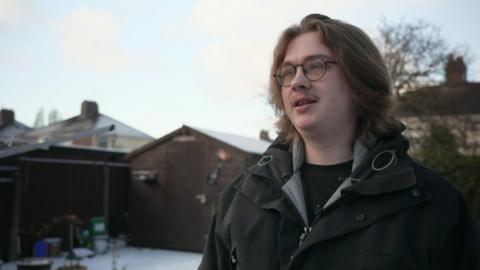 A young man, wearing glasses and a black coat, is mid-conversation, looking to the left of the camera, as he stands in a garden which has snow on the ground