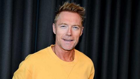 Ronan Keating, wearing a yellow sweatshirt, and standing in front of a black background. He has spiky fair hair and grey stubble; he is smiling at the camera