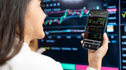 A woman in a white shirt looks at a line graph on her phone, with a big black stock market trading screen in the background.