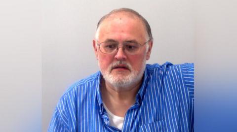 Mark Kerbey wearing glasses and a blue and white striped shirt appearing in his police custody photo.