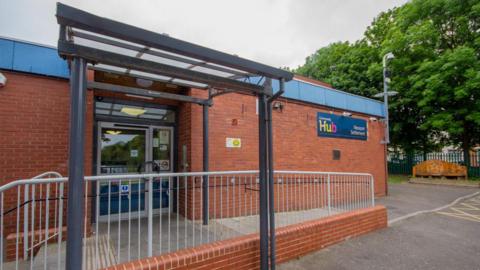 The community hub is a single-storey red brick building with stairs and a ramp leading up to the door.