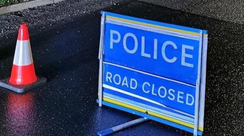 A blue police road closed sign sits on a road. There is also a red and white cone next to it.