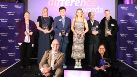 Group of men and women smiling and looking into the camera from a stage, in front of a big purple screen with BBC Radio Sussex and BBC Radio Surrey branding and a Make A Difference awards tv screen.