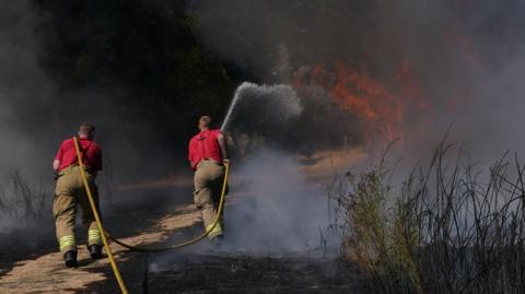 Two firefighters with a yellow hose pipe are directing water on to a grass fire. They are wearing red tops and light coloured trousers and facing the flames.