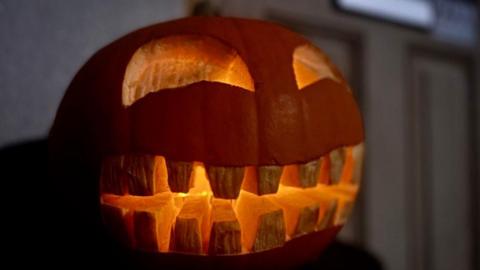 A carved orange pumpkin outside of a light brown door. The pumpkin has semi circle eyes carved and a mouth with lots of rectangular teeth. It is lit up with candles inside 