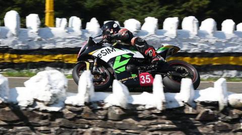 Garry Broughton riding a black motorcycle which has green and white branding on it and the number 35 on a red background. There are white painted stone walls on either side of the course.