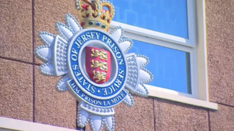 A close up of a sign for Jersey's only prison. The crest is blue with a red shield in the middle with three lions on it. There is also a crown sitting on the blue crest.
