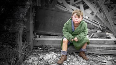 A boy in green clothing looking into the camera surrounded by black-and-white wreckage