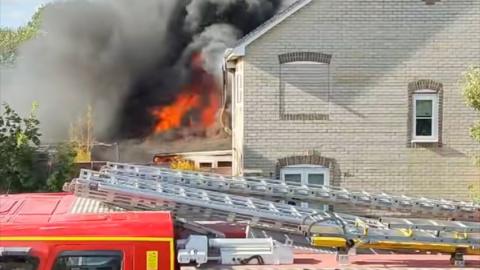 Flames and smoke are leaping up one wall of a grey brick home. The top of a red fire engine and ladders from an arriving fire crew can be seen in the foreground and the sky is obscured by thick black smoke