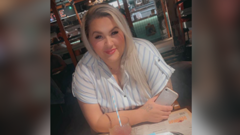 A close up of Sophie sitting at a table with a drink in front of her. It is in a glass with a blue and white straw. Sophie holds a phone in her hand and wears a short sleeved blouse with blue stripes. She has long hair that is blonde and dark.