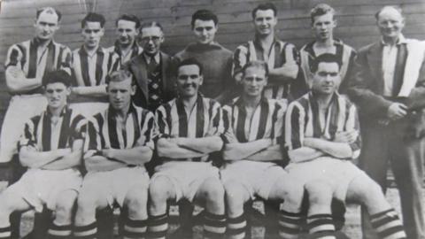 A black and white photo of a football team, its club chairman, and trainer, posing for a team photo. There are five players sitting in the front row, while the other six players, as well as the chairman just left of centre and the trainer on the right, stand behind them. The outfield players wear white-and-dark striped jerseys, with white shorts and hooped socks seemingly of the same colours as the jerseys. The goalkeeper wears a dark top, while the chairman, who has glasses, wears a suit, tie and a patterned waistcoat. The trainer on the right wears a dark jacket and trousers, a white shirt with no tie, and has a towel over his left shoulder.