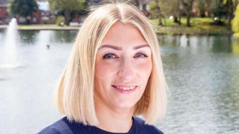 Lorraine is posing for a photo in the sunshine infront of a lake. She has short blonde hair and blue eyes.