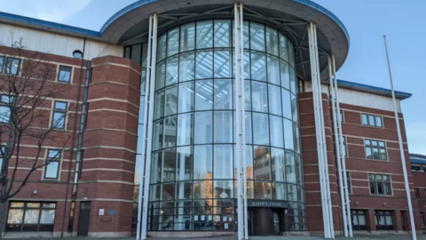 Front view of a modern red-brick justice centre building with a large curved glass atrium entrance, supported by tall white columns and topped with a sweeping roof canopy. The building spans several storeys, with rows of windows on either side of the central glass façade under a clear daylight sky.
