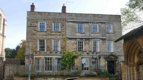 A three-storey pale stone building with panelled window frames and railings outside the property, bordering a path.
