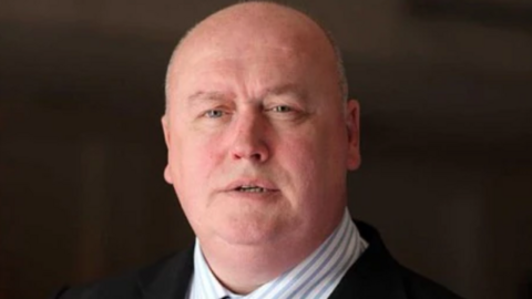 Bald-headed Malcolm Macaskill wearing a dark suit and blue and white striped shirt