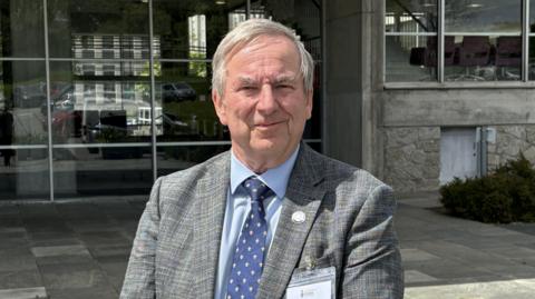 A man with grey hair in a grey jacket with a blue shirt and dark blue tie standing in front of County Hall in Truro