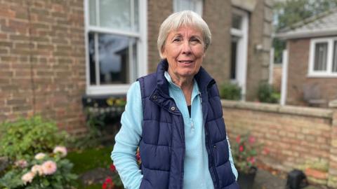 Jenny Pennington wearing a blue shirt and bodywarmer standing with her hands in her pocket in front of her house