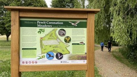 A sign at one of the entrances to the site with a family walking nearby