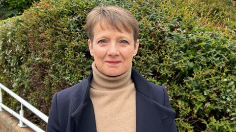 Clare Moody, wearing a brown top and navy jacket, looks ahead and smiles. She has short brown hair