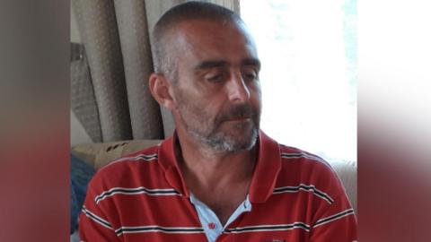 A man is sat by a window, he has a greying beard and short cropped hair. He is wearing a red stripe polo shirt and is looking down away from he camera. 