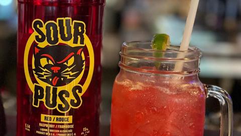 An image showing a close-up of a red Sour Puss bottle, with a black and yellow logo that features a drawing of a cat sticking its tongue out. Next to the bottle is a red cocktail in a mason jar glass with a plastic straw and a lime.