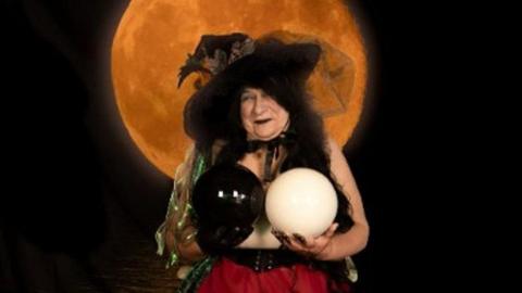 Ros Reynolds holding two crystal balls, one black and one white, to cover the top half of her naked body. She has a red skirt on and witches hat and is posing in front of a dark background with a moon.