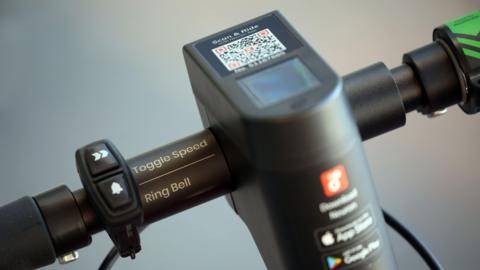 A close-up of a scooter's handlebars. A QR code is in the centre with the words 'Scan & Ride' and a logo advertising the Neuron app.