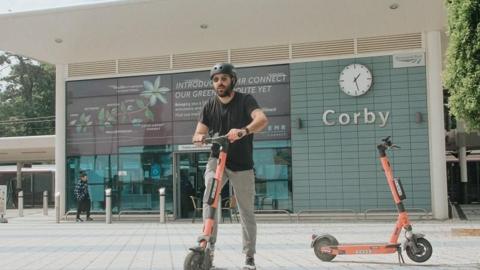 A man with a dark beard wearing a black T-shirt and grey tracksuit trousers rides an orange e-scooter outside Corby railway station. Another orange e-scooter is standing to his left.