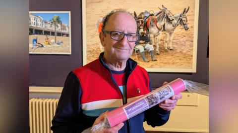 Photographer JJ Waller at his Beside the Sea photography exhibition at the Hastings Museum and Art Gallery. JJ is wearing a striped Fila jacket and is holding a circular tube which says "Greetings from JJ Waller's St Leonards".