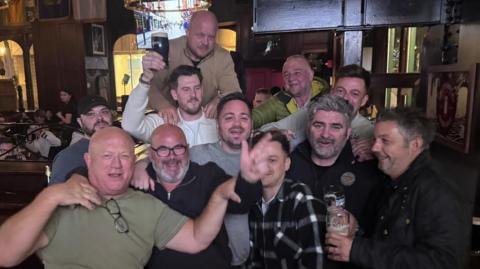 A group of middle-aged men at a pub, some carrying pints of Guinness and looking merry