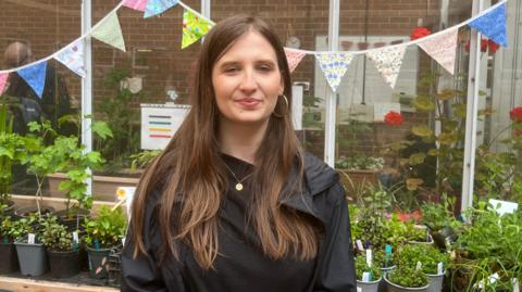 A woman with long brown hair wearing a black T-shirt with black waterproof jacket over the top. She is standing in front of a greenhouse with patterned bunting hanging on it. There are potted plants behind her. She also has a small gold necklace and gold hooped earrings.