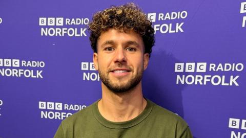 Emiliano Marcondes stands in front of a BBC Radio Norfolk backdrop. He's wearing a green T-shirt and is smiling