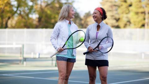 A stock image of two women chatting on a tennis court. On the left a woman with blonde hair, a white sports jacket and a blue top. On the right a woman with brown hair, a white sports jacket and a red headband. 