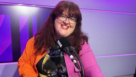 A smiling woman with long brown hair and glasses wearing a colourful orange-and-pink jacket in a recording studio