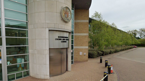Google Street View of the front of Bournemouth Crown Court. The revolving doors are closed and a sign in black writing above the door says Bournemouth Crown and County Courts. Above the sign on the round entranceway is a round crest with a central badge flanked by a lion and a horse.