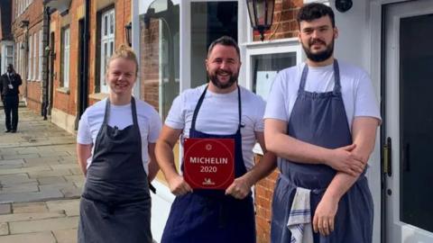 Paul Foster pictured in the middle of a smiling blonde woman and a tall man with black hair and a full beard. Paul is smiling while holding a red plaque which reads "Micheline 2020". All three are wearing black trousers, a white t-shirt and dark blue demin apron.