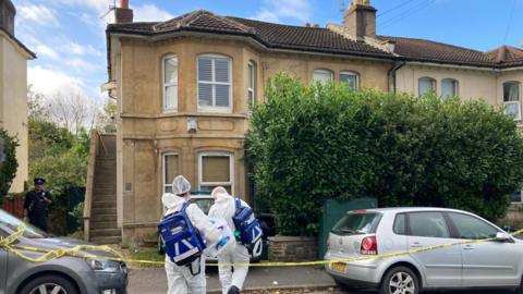 Two forensic examiners entering a house in Bristol wearing white suits, blue latex gloves and hairnets. They are carrying large blue backpacks. On the street there are two parked cars with yellow police tape wrapped around their wing mirrors to make a police cordon. The house is a sand coloured two storey building with bay windows and a staircase leading up to the front door on the left. 