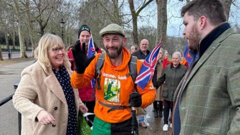 A man in an orange top carrying a rucksack and wearing a flat cap. He is smiling and a woman to his left in a cream coat is also smiling. Several other people are behind them. 