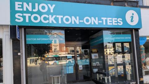 A tourist information building with "Enjoy Stockton-on-Tees" written on a large light blue sign over the door. The windows are glass with electric automatic doors in the centre.