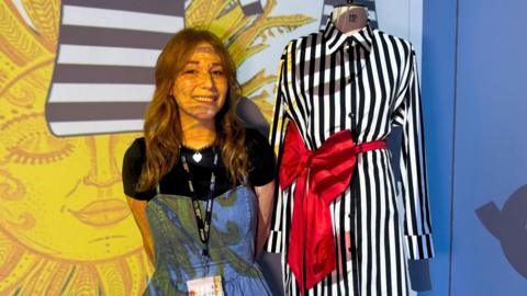 A woman stands beside a mannequin dressed in a black‑and‑white striped shirt dress with a large red bow, set against a bright yellow sun illustration projected on the wall. 