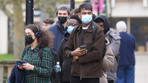 A group of people stand in a queue, most are wearing masks over their mouths and nose and the woman and man at the front are looking at their phones